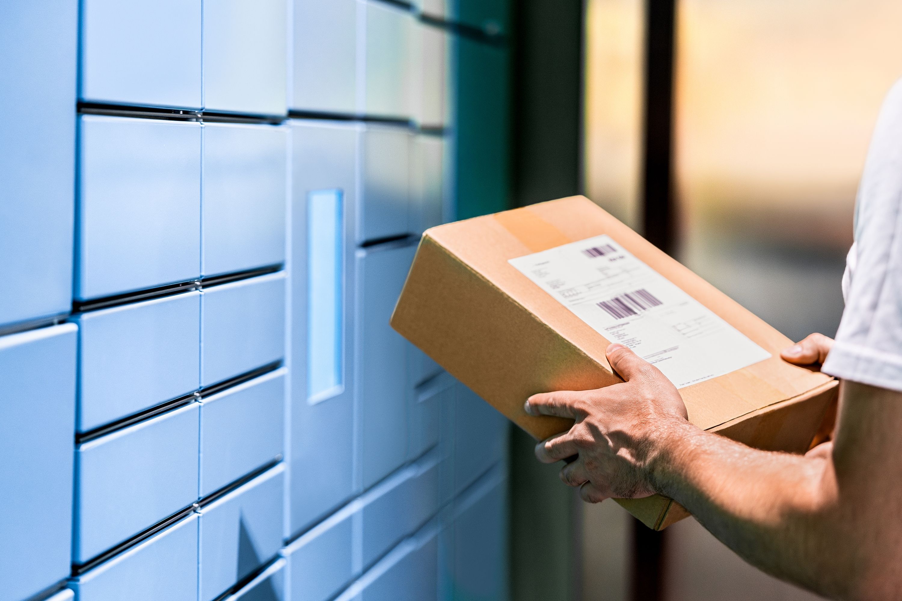 Secure package mailbox person holding a small brown box in front of blue package mailboxes